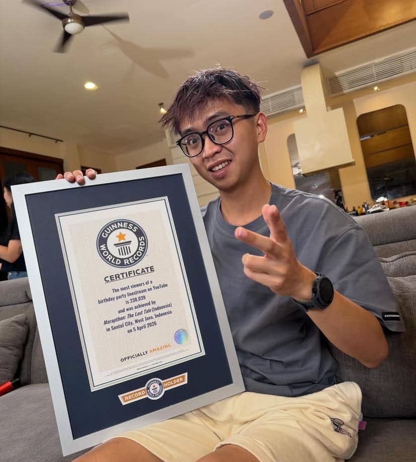 Seorang pria muda berkacamata duduk di sofa sambil memegang sertifikat Guinness World Records dengan ekspresi bangga di ruang tamu.