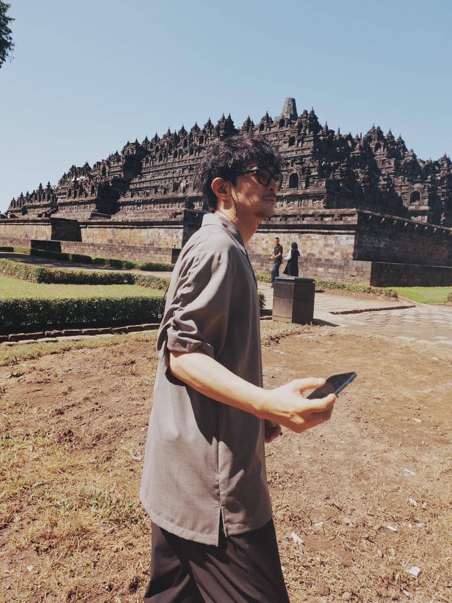 Seorang pria berjalan sambil memegang ponsel di area Candi Borobudur dengan langit cerah dan suasana siang hari yang tenang.