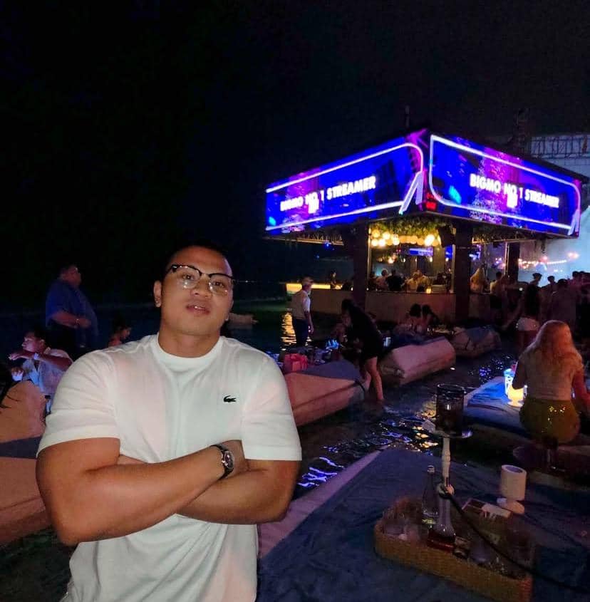 Seorang pria berkaus putih berpose dengan tangan terlipat di area pesta malam hari dengan pencahayaan neon dan keramaian di sekitarnya.