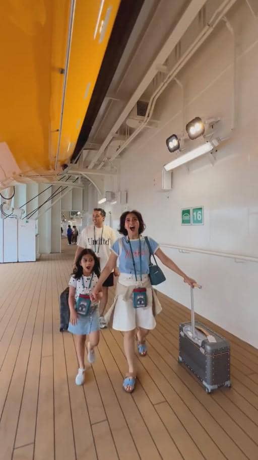 Seorang wanita bersama anak kecil dan seorang pria berjalan di dek kapal pesiar dengan koper dan tas, tampak bersemangat menikmati perjalanan.