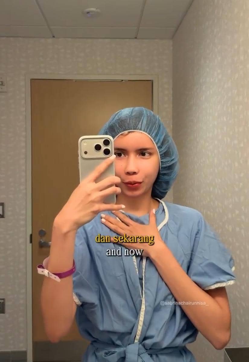 Seorang perempuan mengenakan pakaian medis biru dan penutup kepala sedang berfoto selfie di depan cermin di sebuah ruangan rumah sakit.