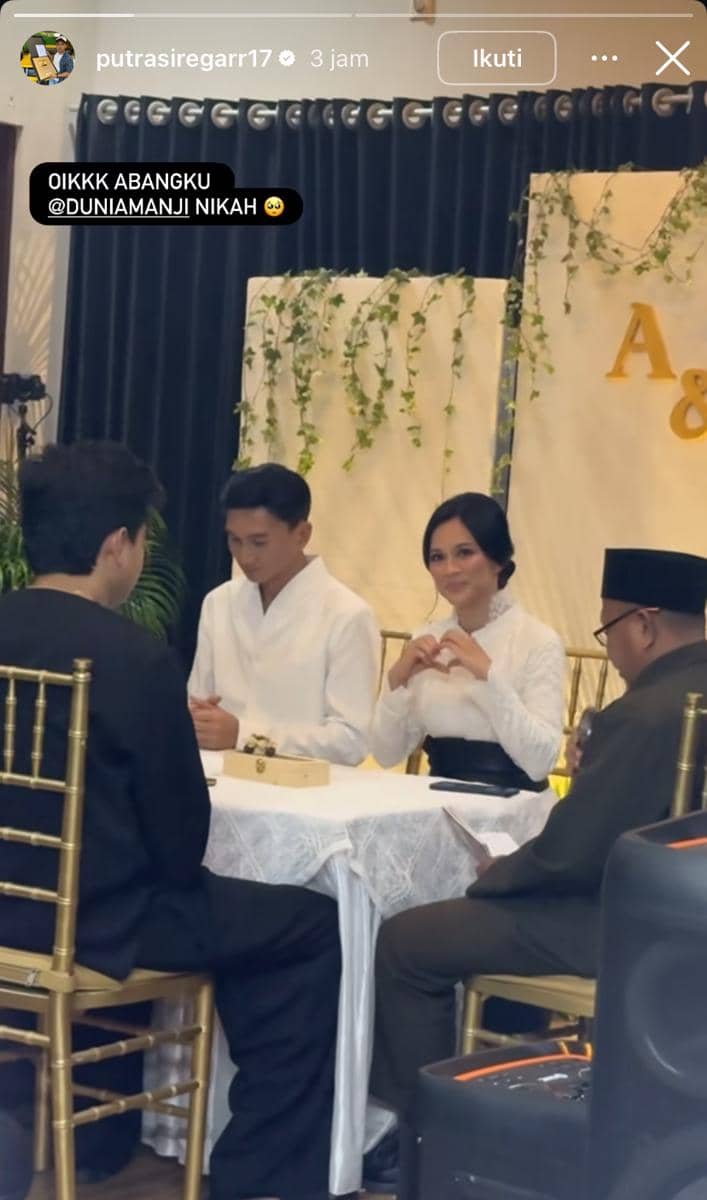 Pasangan pengantin mengenakan busana putih duduk di meja akad nikah dengan dekorasi daun hijau dan inisial nama di latar belakang.