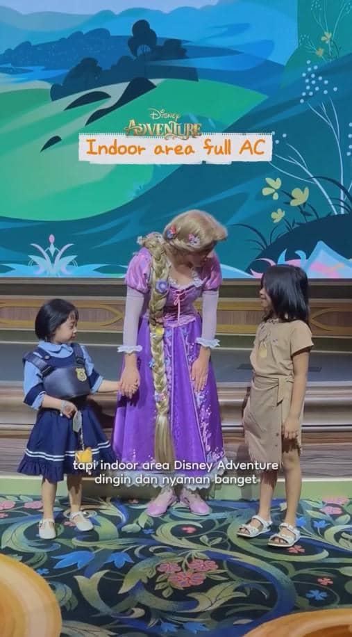 Seorang karakter putri Disney bergaun ungu berbicara dengan dua anak kecil di area indoor bertema Disney Adventure yang berwarna cerah.