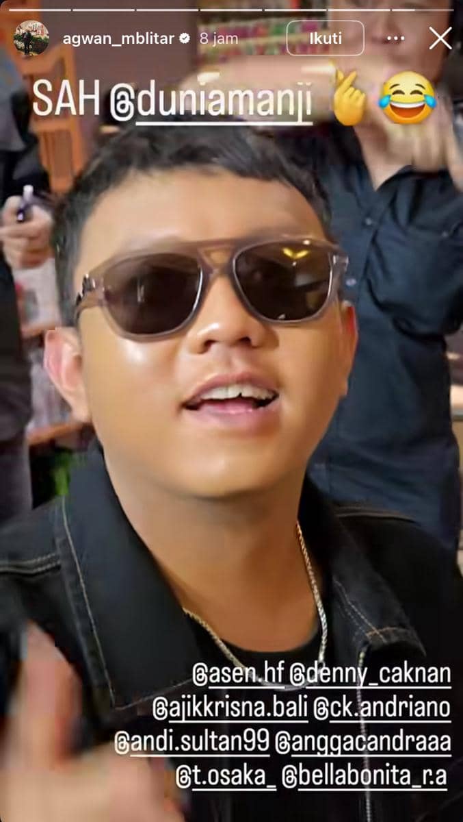 Seorang pria mengenakan kacamata hitam dan jaket hitam tersenyum di acara pernikahan Anji dan Desy dengan suasana meriah di sekitarnya.