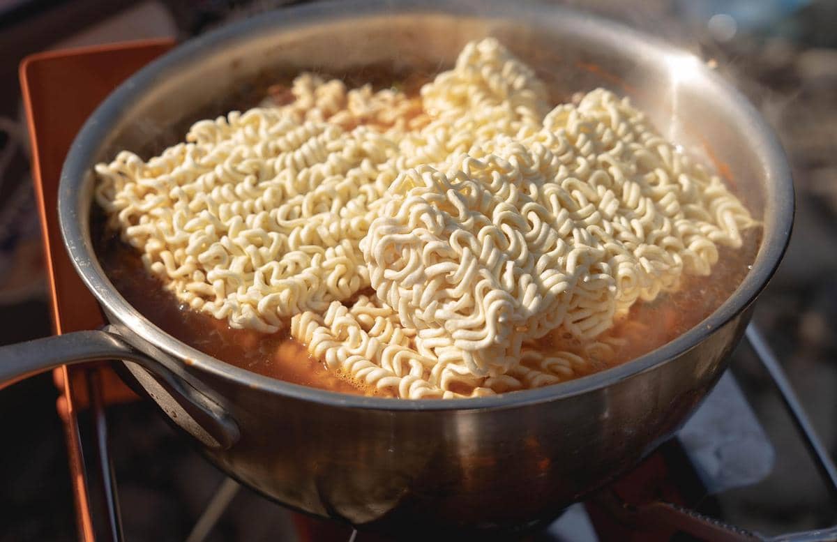 Panci berisi air mendidih dengan beberapa blok mi instan ramen yang sedang direbus di atas kompor luar ruangan.