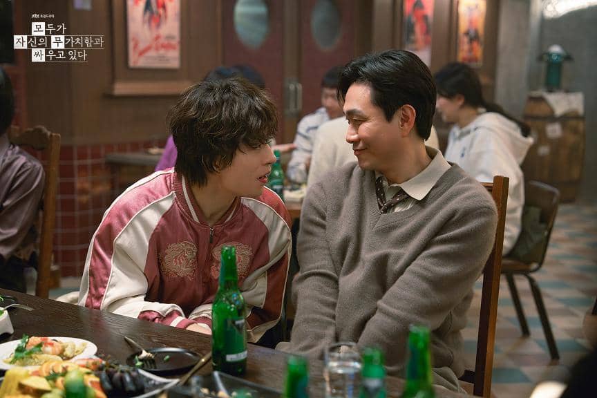 Dua karakter dalam drama Korea We Are All Trying Here duduk berhadapan di meja makan restoran, saling tersenyum hangat di tengah suasana akrab.