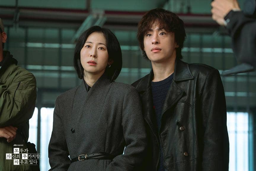 Dua karakter utama dalam drama Korea We Are All Trying Here berdiri berdampingan dengan ekspresi serius di sebuah ruangan.