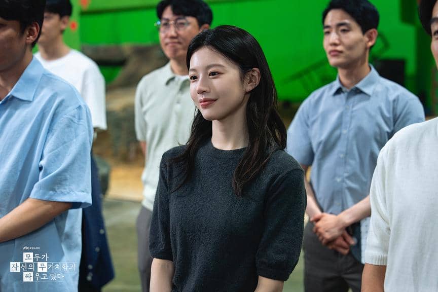 Seorang wanita berambut hitam mengenakan kaus abu-abu berdiri di antara beberapa orang pria dalam adegan drama Korea We Are All Trying Here.