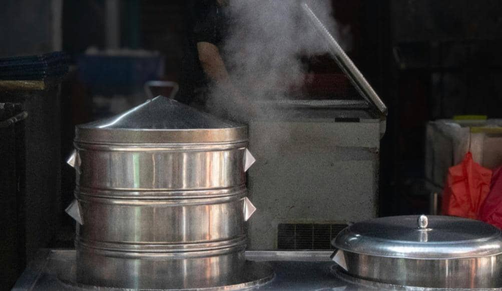 Panci pengukus besar dari stainless steel mengeluarkan uap panas di dapur, menunjukkan proses memasak dengan cara mengukus.