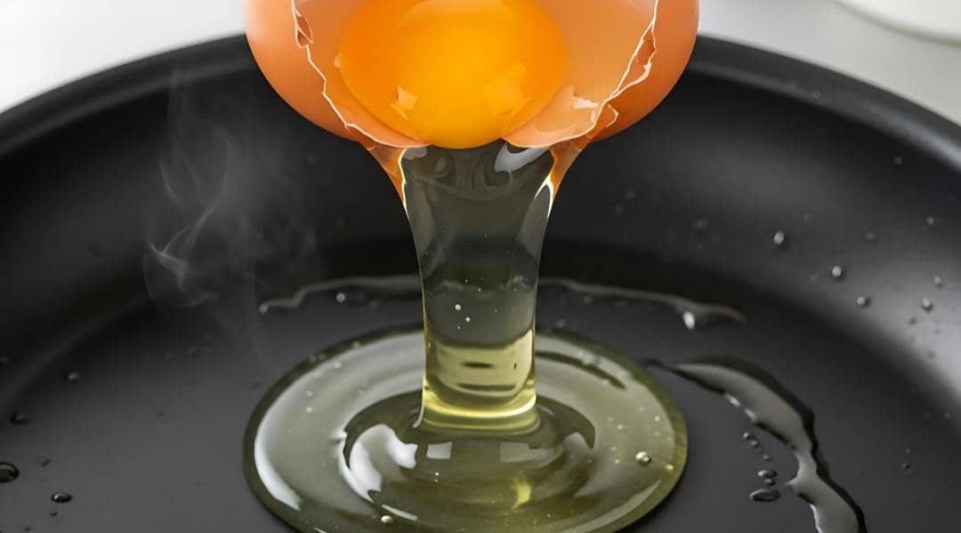 Telur dipecahkan dengan kuning telur dan putih telur mengalir ke dalam wajan hitam, memperlihatkan proses pemisahan putih telur.