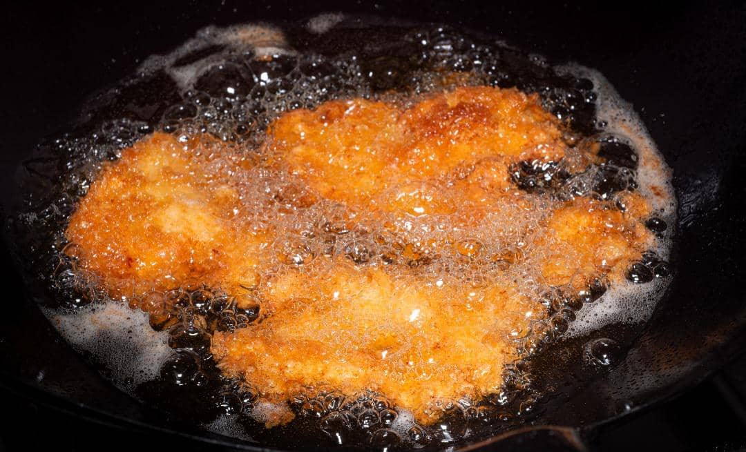 Beberapa potong nugget berwarna keemasan sedang digoreng dalam minyak panas di wajan dengan gelembung minyak yang banyak.