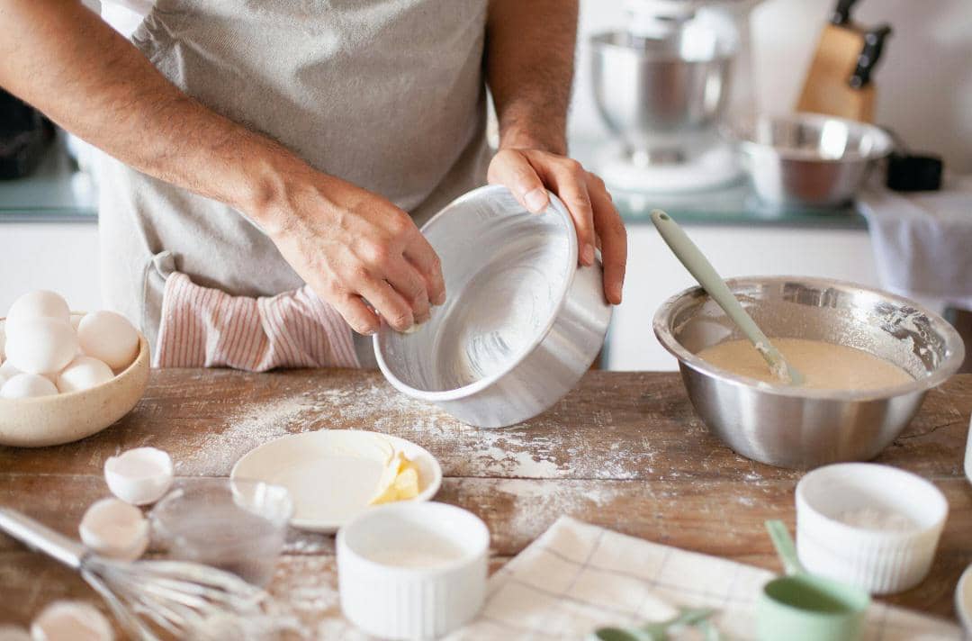 Seseorang mengenakan celemek sedang mengoles loyang dengan margarin di dapur, di meja terdapat adonan, telur, dan peralatan memanggang.