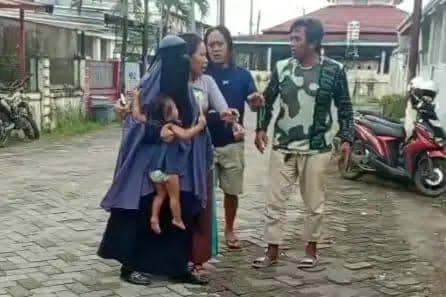 Seorang pria berbicara dengan seorang wanita yang menggendong anak kecil di jalan perumahan, disaksikan oleh seorang pria lain di dekat mereka.