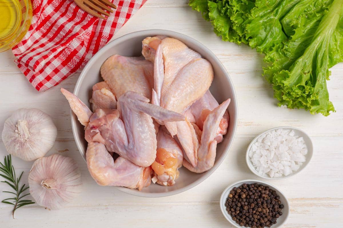 Potongan daging ayam mentah dalam mangkuk putih dikelilingi bawang putih, garam, lada hitam, daun selada, dan minyak zaitun di atas meja kayu.