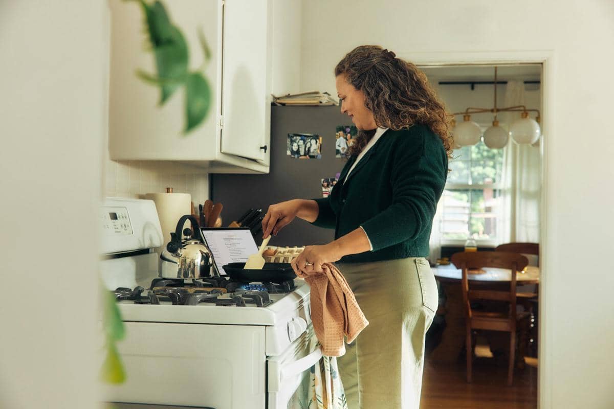 Seorang wanita sedang memasak di dapur dengan api kecil sambil melihat resep di laptop di atas kompor gas.
