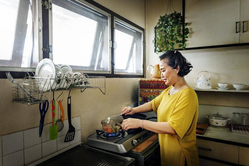 Seorang perempuan mengenakan baju kuning sedang memasak daging di dapur rumah dengan peralatan masak dan piring tersusun rapi di rak.