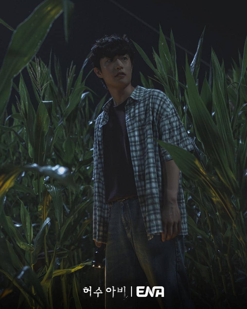Seorang pria berdiri di tengah ladang jagung pada malam hari dengan ekspresi waspada, mengenakan kemeja kotak dan membawa senter kecil.