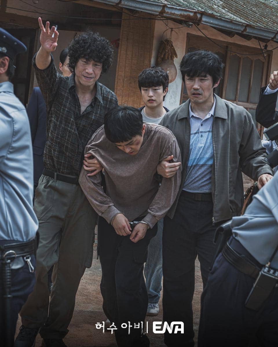 Beberapa pria membantu seseorang yang tampak lemah berjalan di antara petugas polisi dalam adegan drama The Scarecrow.