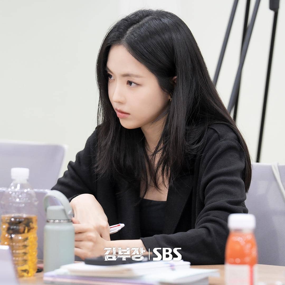 Seorang perempuan berambut hitam panjang mengenakan pakaian hitam sedang duduk serius di meja dengan botol minuman dan dokumen di depannya.