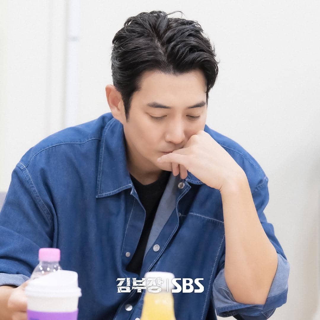 Seorang pria mengenakan kemeja denim biru duduk sambil menatap ke bawah dengan ekspresi serius di meja dengan minuman di depannya.