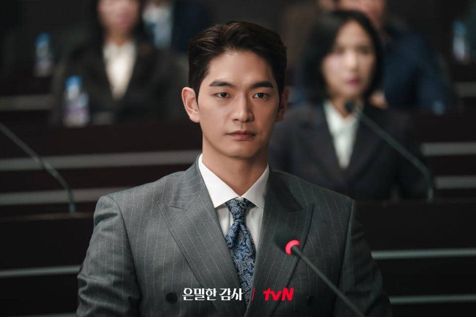 Seorang pria mengenakan setelan jas abu-abu dan dasi biru duduk di ruang sidang dengan mikrofon di depannya dalam cuplikan drama Filing for Love.