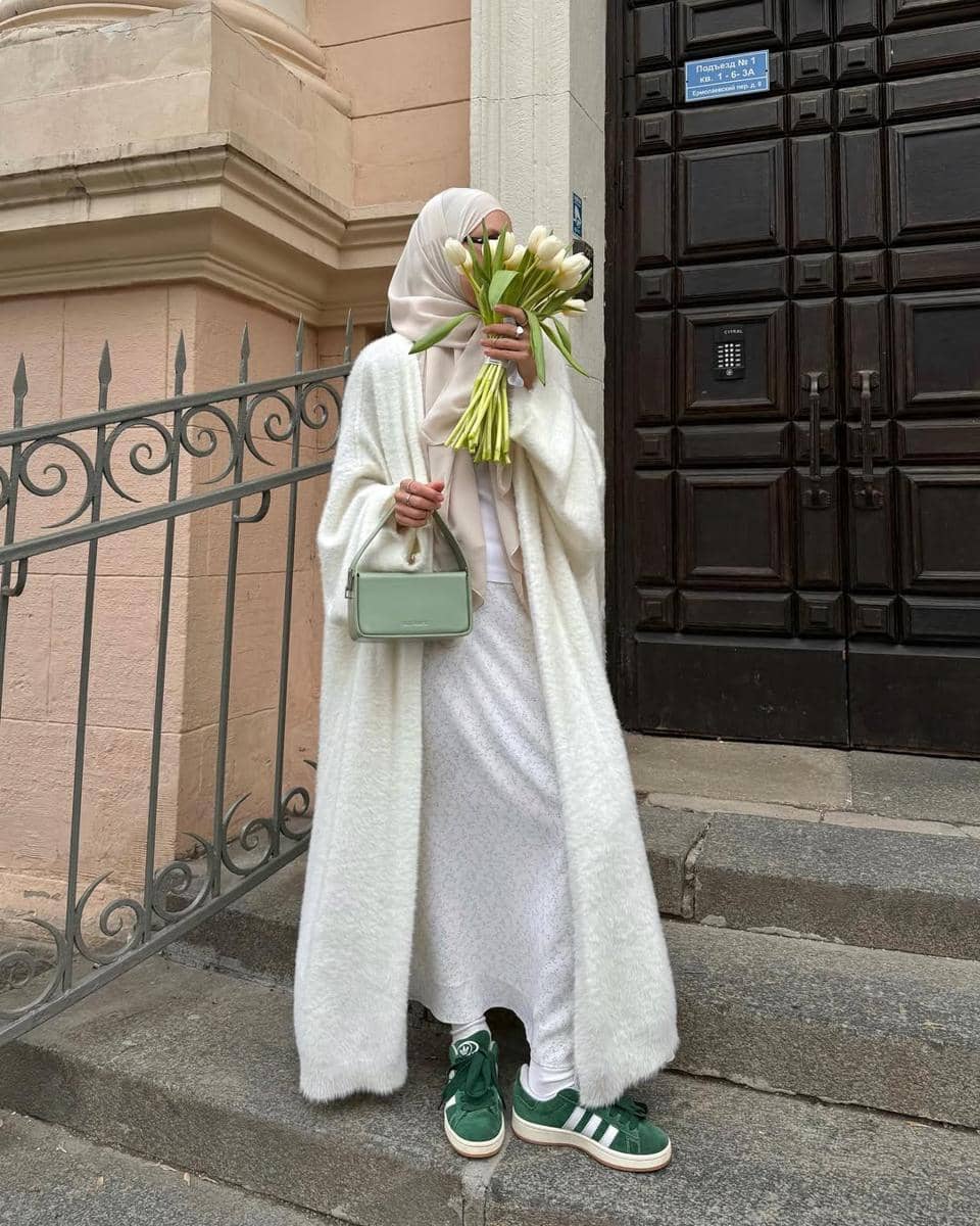 Seorang wanita berhijab mengenakan long coat putih, memegang buket bunga tulip putih, dan tas kecil hijau muda di depan pintu besar kayu.