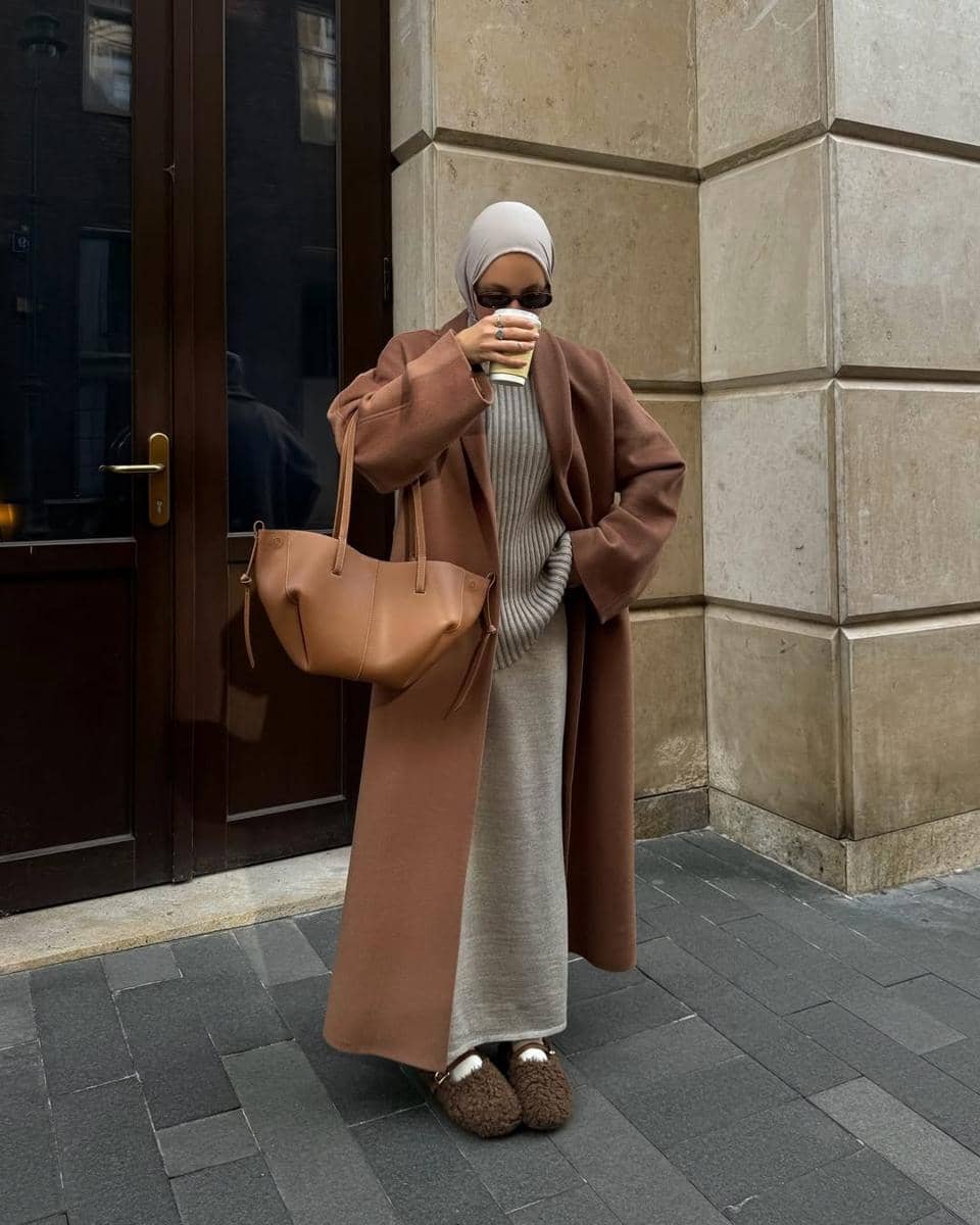 Seorang wanita berhijab mengenakan long coat cokelat, membawa tas besar warna senada, dan memegang kopi di depan bangunan batu.