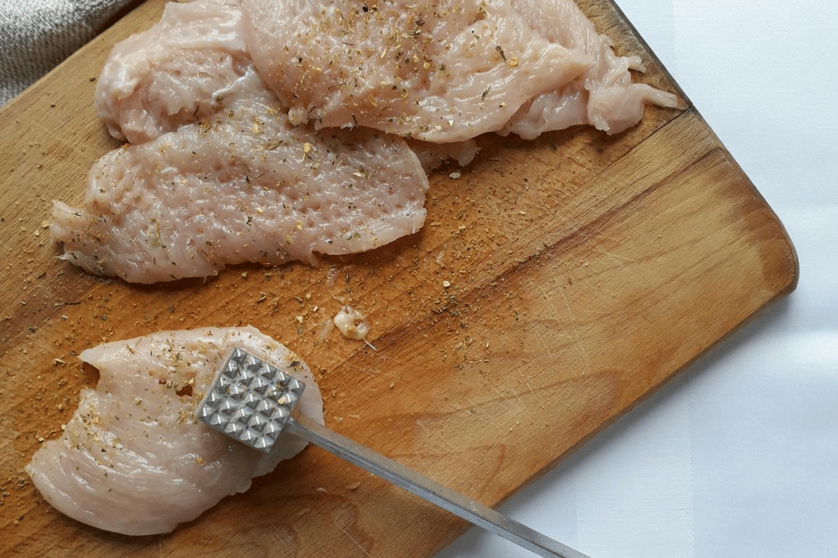 Potongan daging ayam mentah dibumbui rempah di atas talenan kayu dengan pemukul daging logam untuk persiapan ayam crispy.
