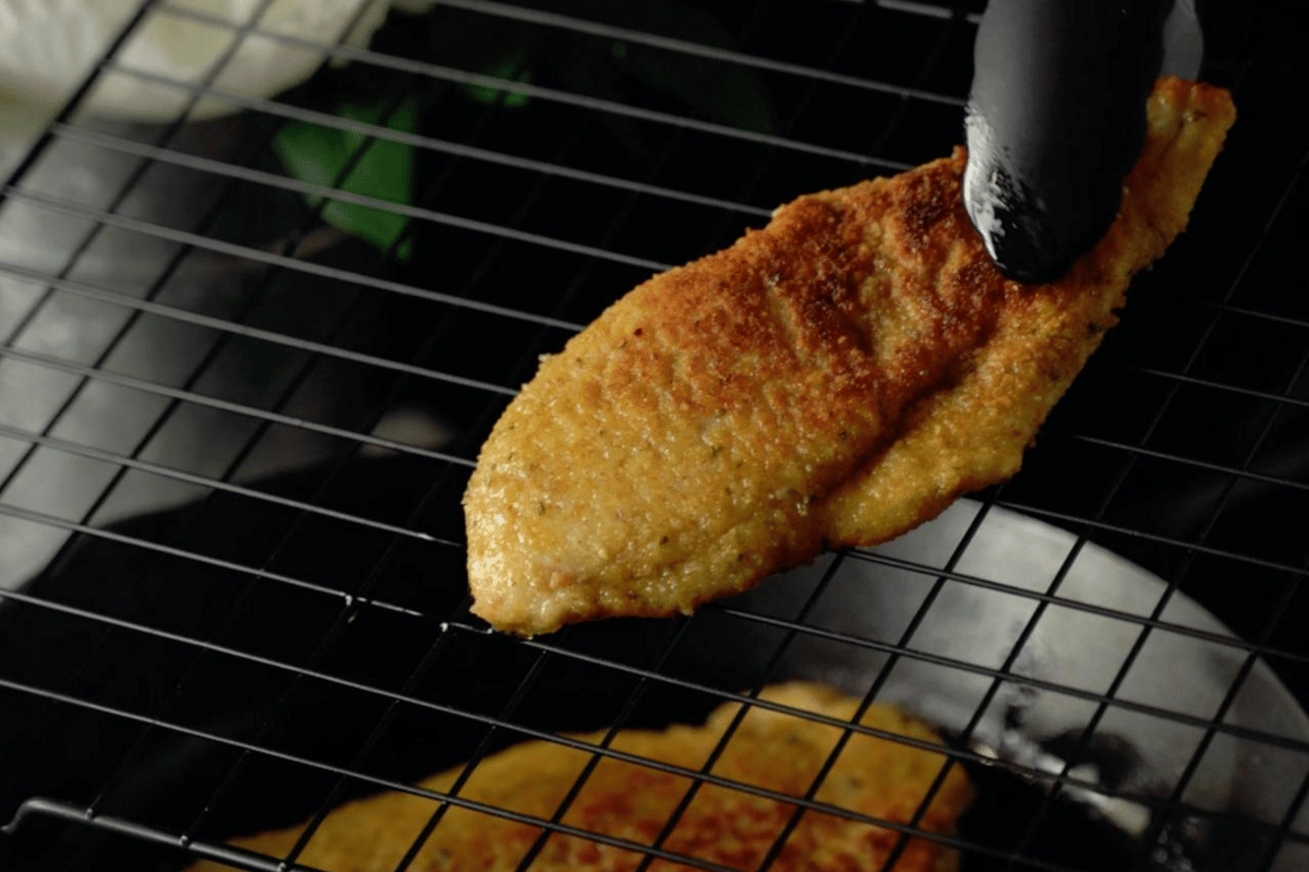Potongan ayam crispy ala Milan berwarna keemasan diangkat dengan penjepit hitam di atas rak pendingin logam.