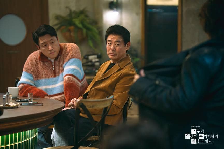 Dua pria duduk di bar dengan ekspresi serius dalam adegan drama Korea We Are All Trying Here, suasana hangat dengan pencahayaan lembut.