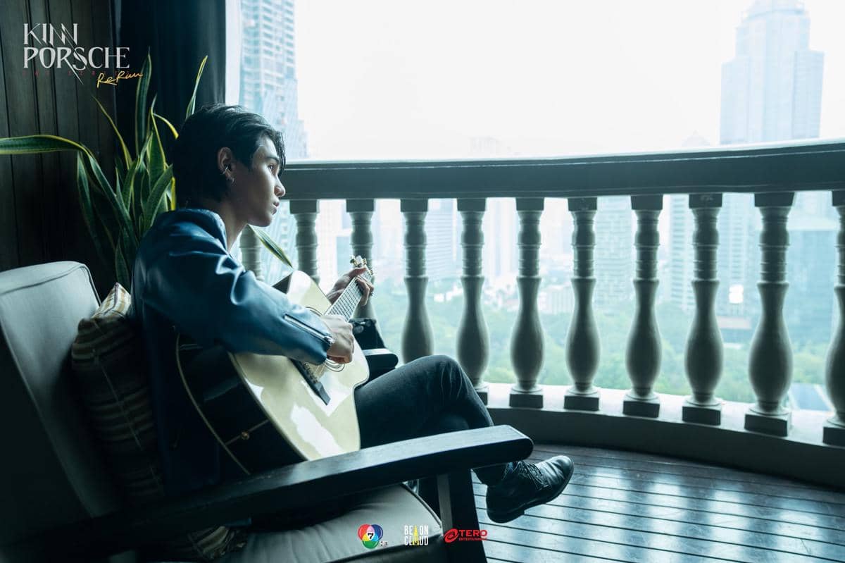 Seorang pria duduk di balkon gedung tinggi sambil memainkan gitar akustik, mengenakan jas biru dan menatap ke arah luar jendela.
