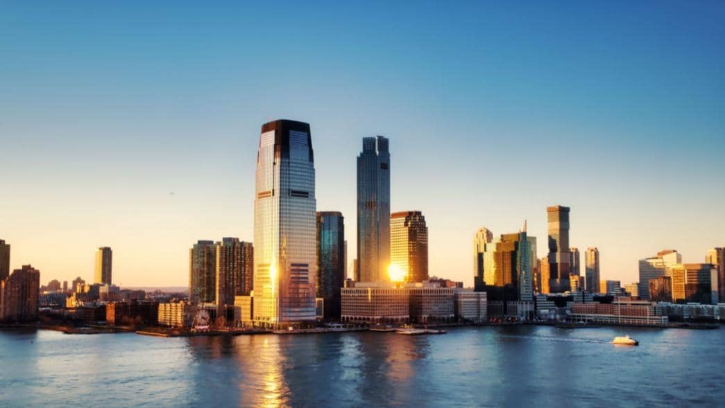 Pemandangan gedung pencakar langit di Jersey City saat matahari terbenam dengan pantulan cahaya di permukaan air sungai.