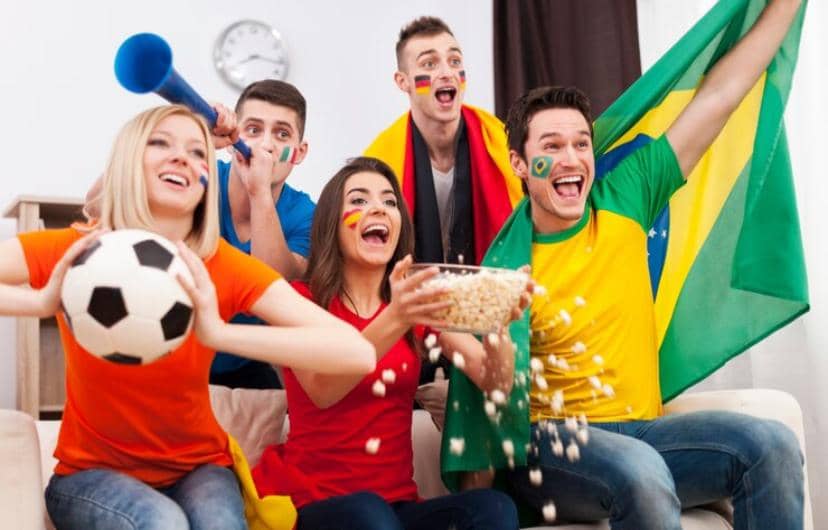 Sekelompok teman bersorak gembira sambil menonton pertandingan sepak bola, mengenakan baju dan bendera negara peserta Piala Dunia.
