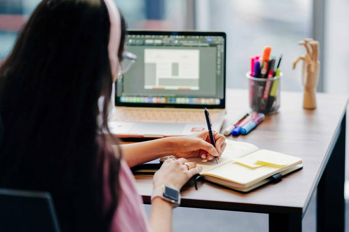 Seseorang menulis di buku catatan sambil belajar desain di laptop dengan alat tulis warna-warni di meja kerja.