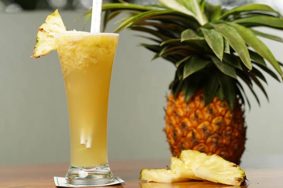 Segelas jus nanas segar berwarna kuning dengan potongan nanas di tepi gelas, disajikan di atas meja bersama buah nanas utuh di belakangnya.