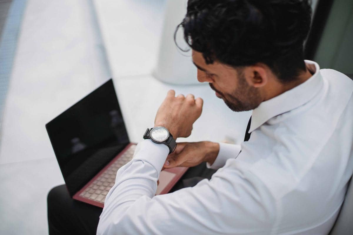 Seorang pria mengenakan kemeja putih duduk di depan laptop sambil melihat jam tangan di pergelangan tangannya.