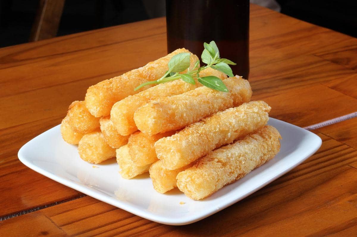 Tumpukan crabstick goreng crispy berwarna keemasan di atas piring putih dengan hiasan daun hijau di meja kayu.