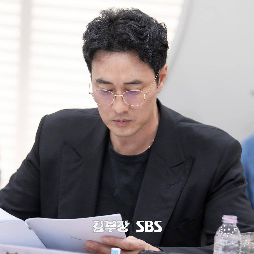 Seorang aktor mengenakan jas hitam dan kacamata bulat sedang membaca naskah di meja dalam sesi pembacaan naskah drama Agent Kim Reactivated.