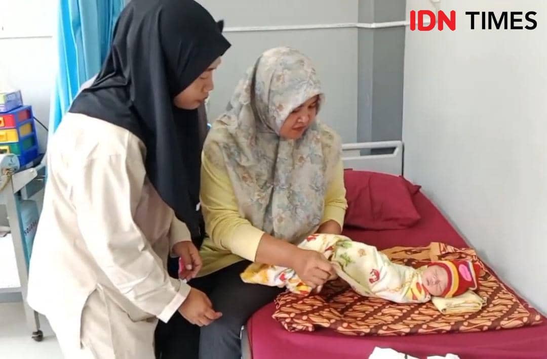 Dua perempuan berhijab sedang merawat bayi yang dibedong di atas tempat tidur rumah sakit dengan selimut bermotif warna-warni.