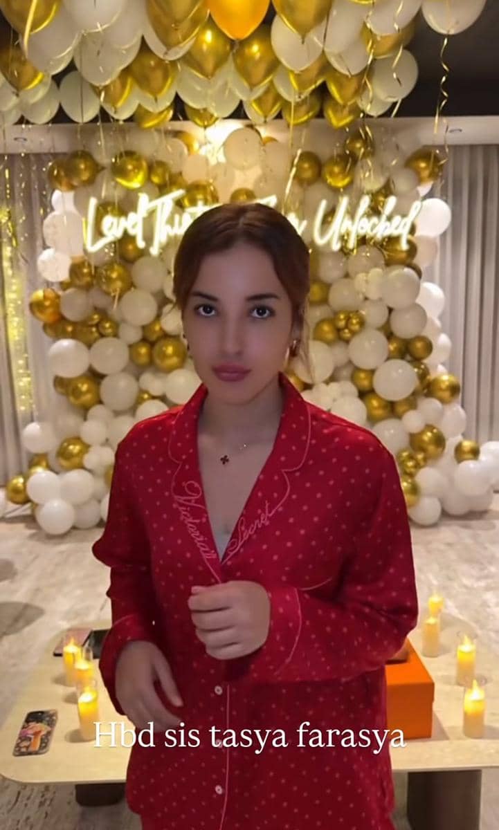 Seorang wanita mengenakan piyama merah berdiri di depan dekorasi balon putih dan emas dengan tulisan neon di latar belakang.