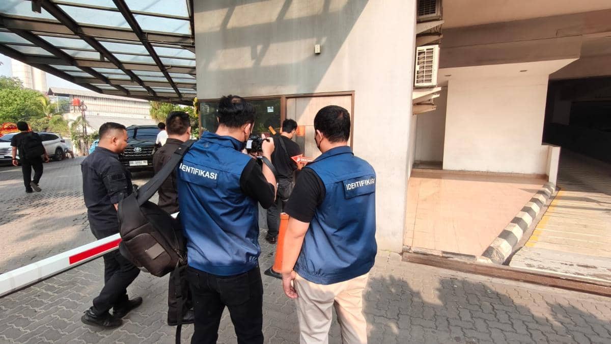 Petugas identifikasi mengenakan rompi biru sedang melakukan pemeriksaan di area luar Hotel B Fashion, Jakarta Barat pada siang hari.
