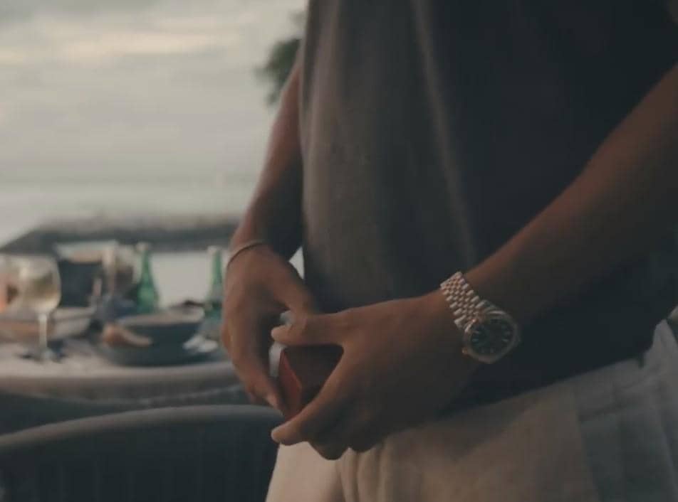 Seorang pria mengenakan jam tangan perak memegang kotak cincin kecil di tepi pantai dengan latar meja makan romantis.