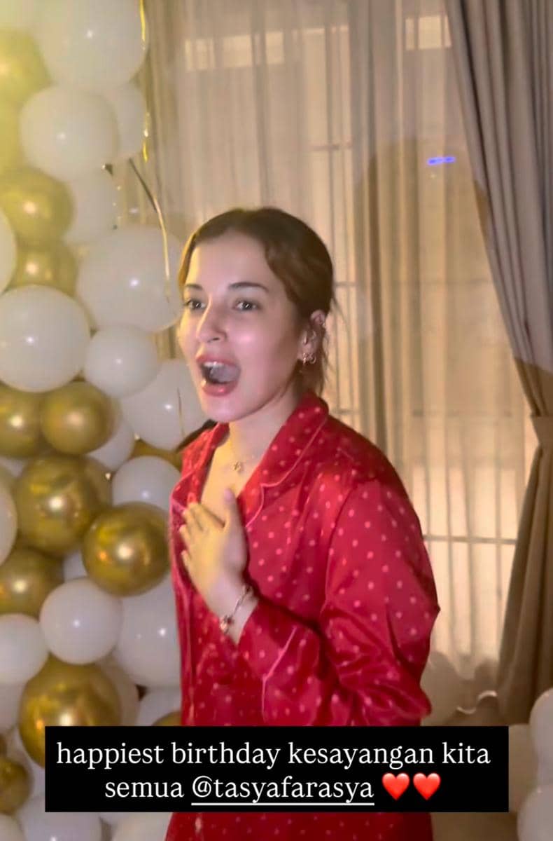 Seorang perempuan mengenakan piyama merah bermotif polkadot tersenyum terkejut di depan dekorasi balon putih dan emas saat perayaan ulang tahun.