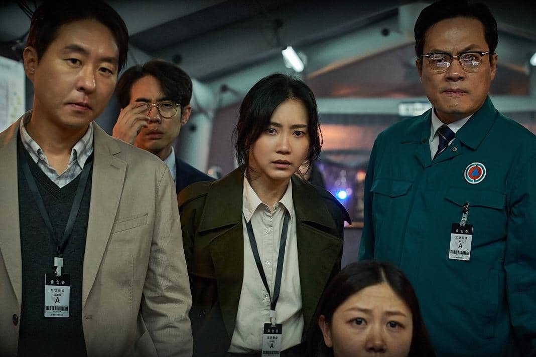 Beberapa pemeran film Korea Colony berdiri dengan ekspresi serius di dalam ruangan, mengenakan pakaian formal dan kartu identitas.