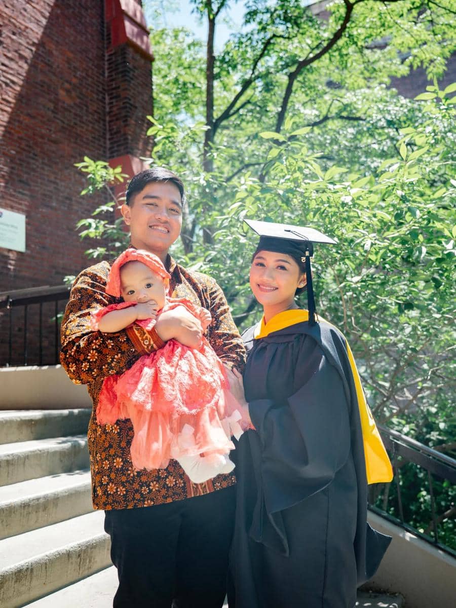 Seorang perempuan mengenakan toga wisuda berfoto bersama pria berbaju batik yang menggendong bayi di luar ruangan dengan latar pepohonan.
