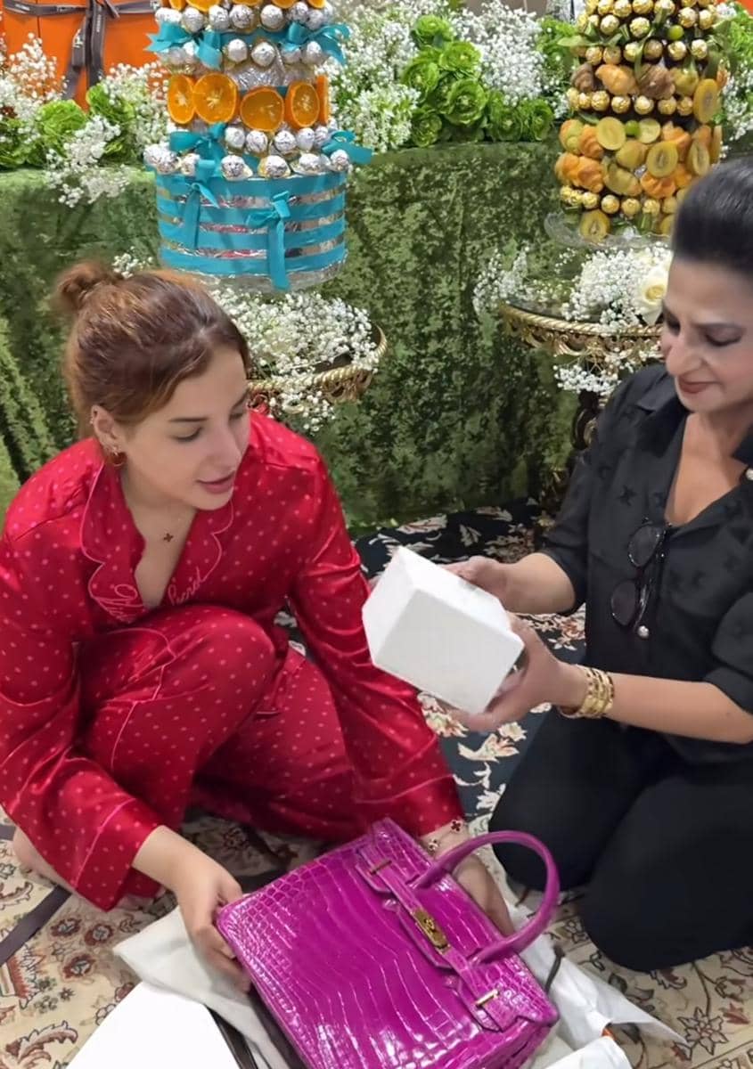 Seorang wanita mengenakan piyama merah duduk di lantai bersama wanita lain yang memegang kotak putih, dengan tas ungu dan dekorasi bunga di sekitarnya.