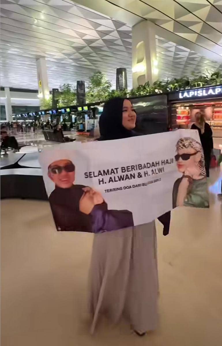 Seorang perempuan berhijab memegang spanduk bertuliskan ucapan selamat beribadah haji di area bandara yang modern dan ramai.