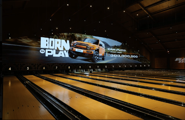 Layar besar di arena bowling menampilkan iklan mobil iCAR V23 berwarna oranye dengan tulisan harga resmi yang diumumkan.
