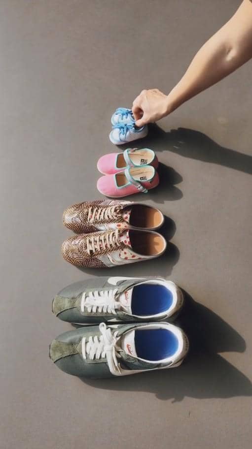 Empat pasang sepatu tersusun berurutan dari besar ke kecil, dengan tangan memegang sepatu bayi biru di bagian atas.