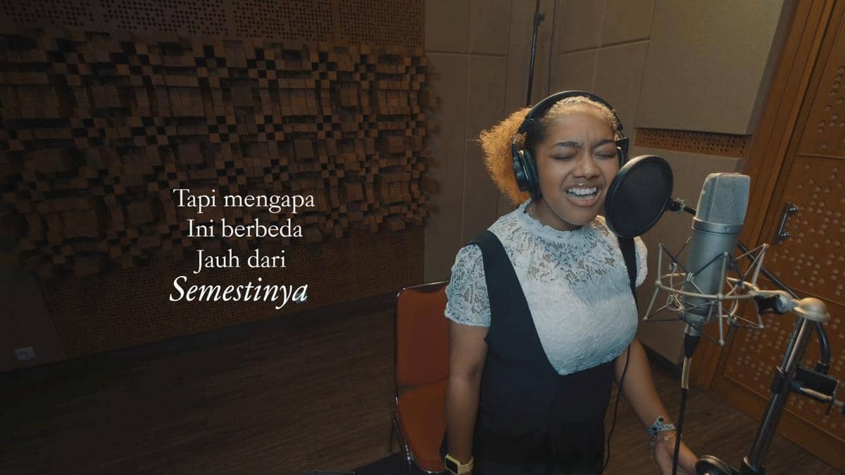Seorang penyanyi sedang bernyanyi dengan penuh ekspresi di depan mikrofon di studio rekaman dengan teks lirik di layar.